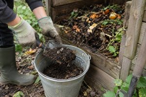 Une personne portant des gants et des bottes utilise une petite pelle pour transférer du compost d'un bac
